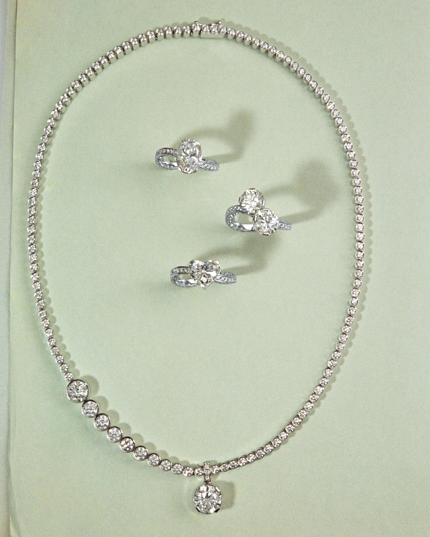 A white gold diamond tennis collier circles around three white gold rings with large center stones placed in a light green background.